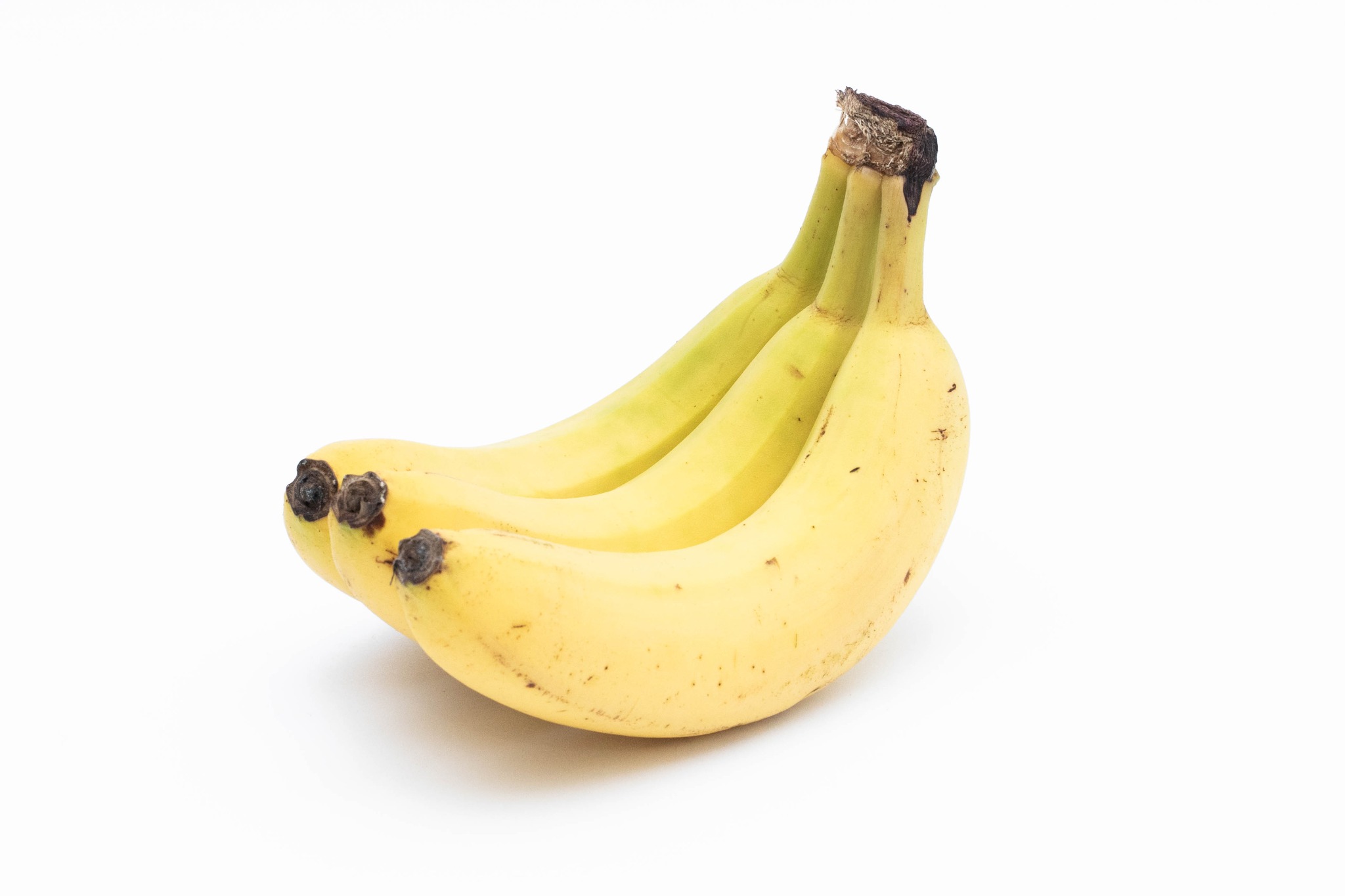 Trzy dojrzałe banany w pęczku, widoczne brązowe plamki na skórce, na białym tle.