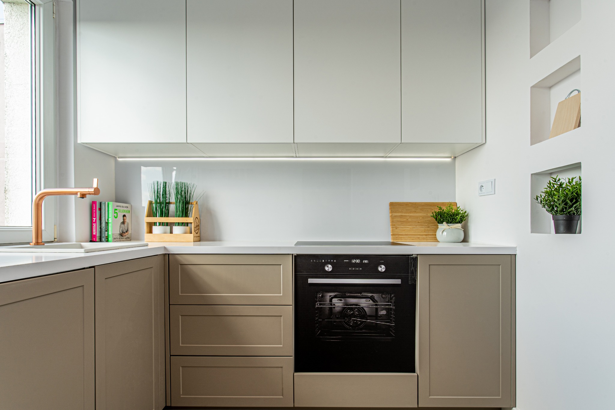 Nowoczesna kuchnia w neutralnych kolorach: szare szafki, białe blaty, czarny piekarnik i miedziana bateria, z akcentami zieleni w postaci roślin doniczkowych i książek kucharskich.