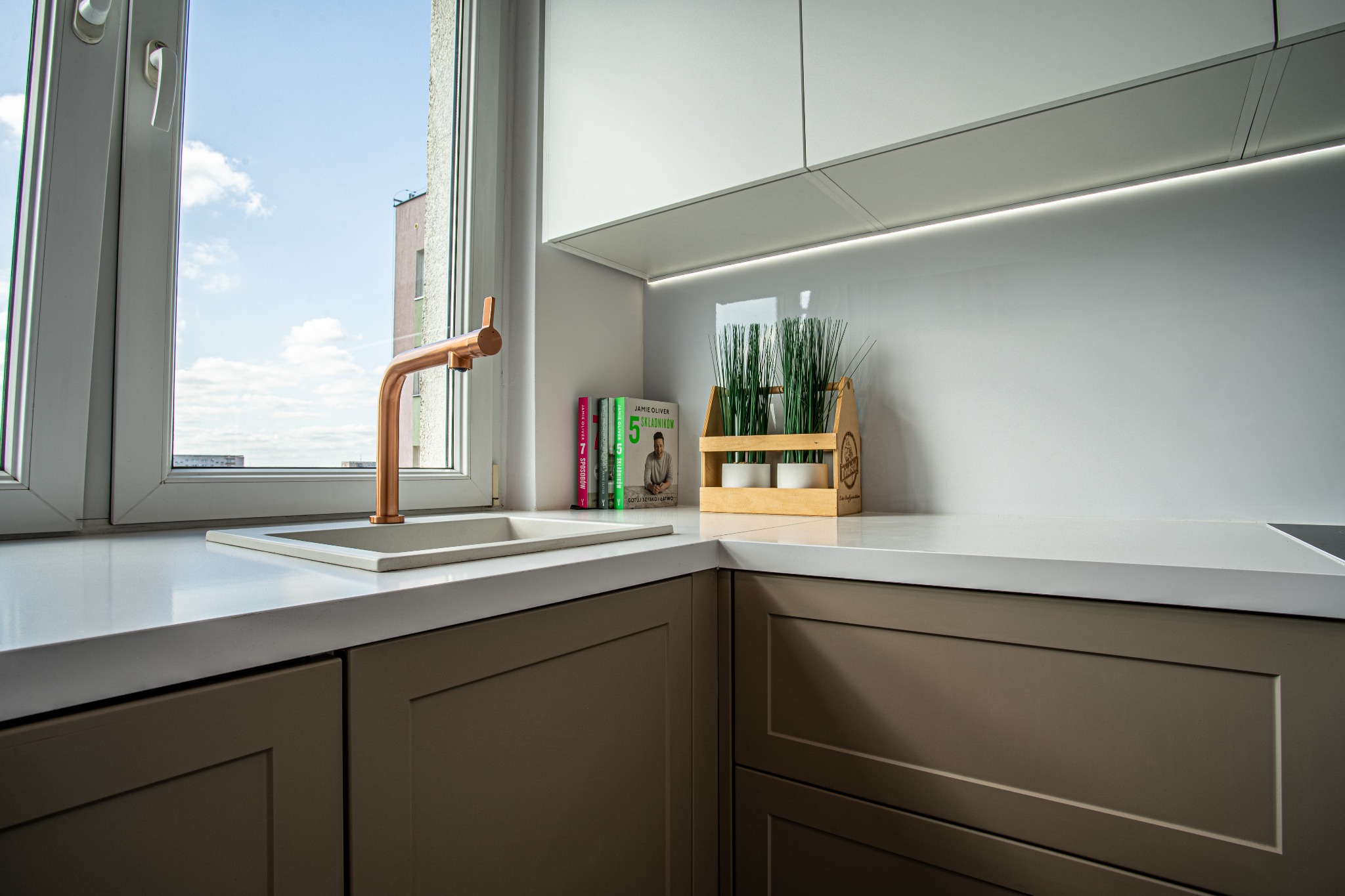 Nowoczesna kuchnia z miedzianą baterią przy oknie, jasnym blatem, szarymi szafkami i ziołami w drewnianej skrzynce obok książek kucharskich Jamiego Olivera.