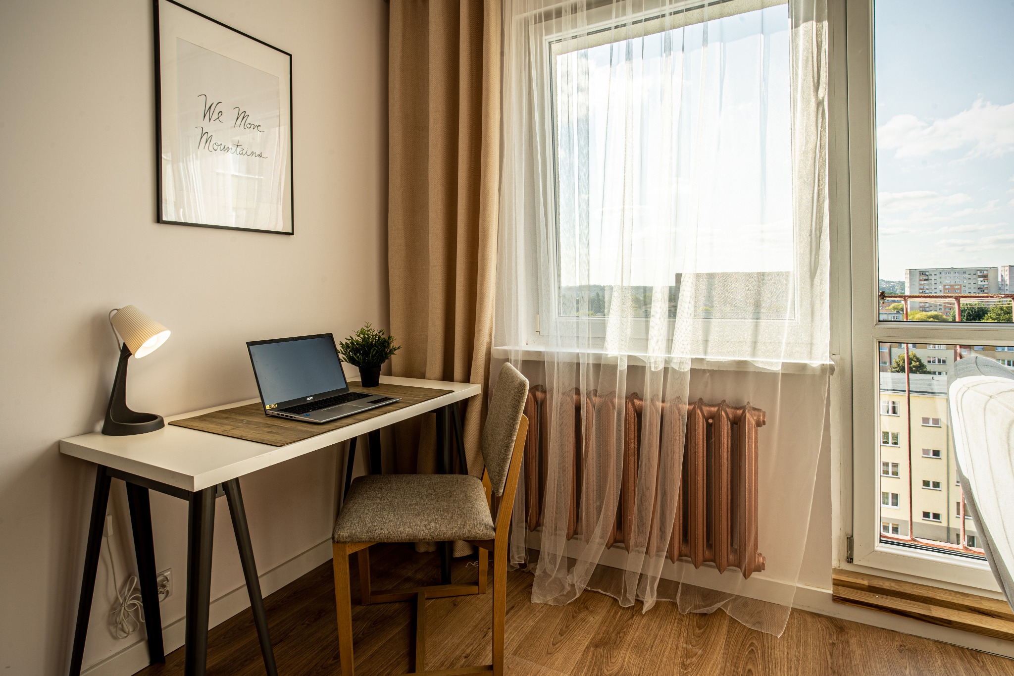 Jasne wnętrze pokoju z biurkiem, laptopem, lampką, krzesłem i widokiem z okna na miasto w słoneczny dzień. Na ścianie plakat z napisem 'We Move Mountains'.