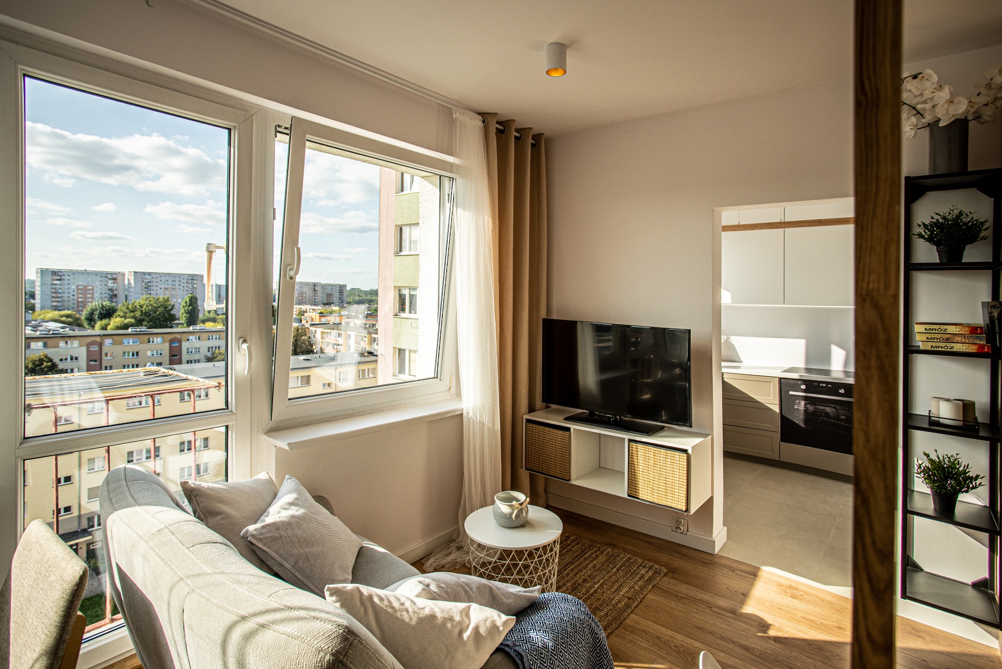 Jasne wnętrze salonu w Szczecinie z dużym oknem, przez które widać panoramę miasta. Wnętrze urządzone w nowoczesnym stylu, z szarą sofą, telewizorem i regałem na książki.