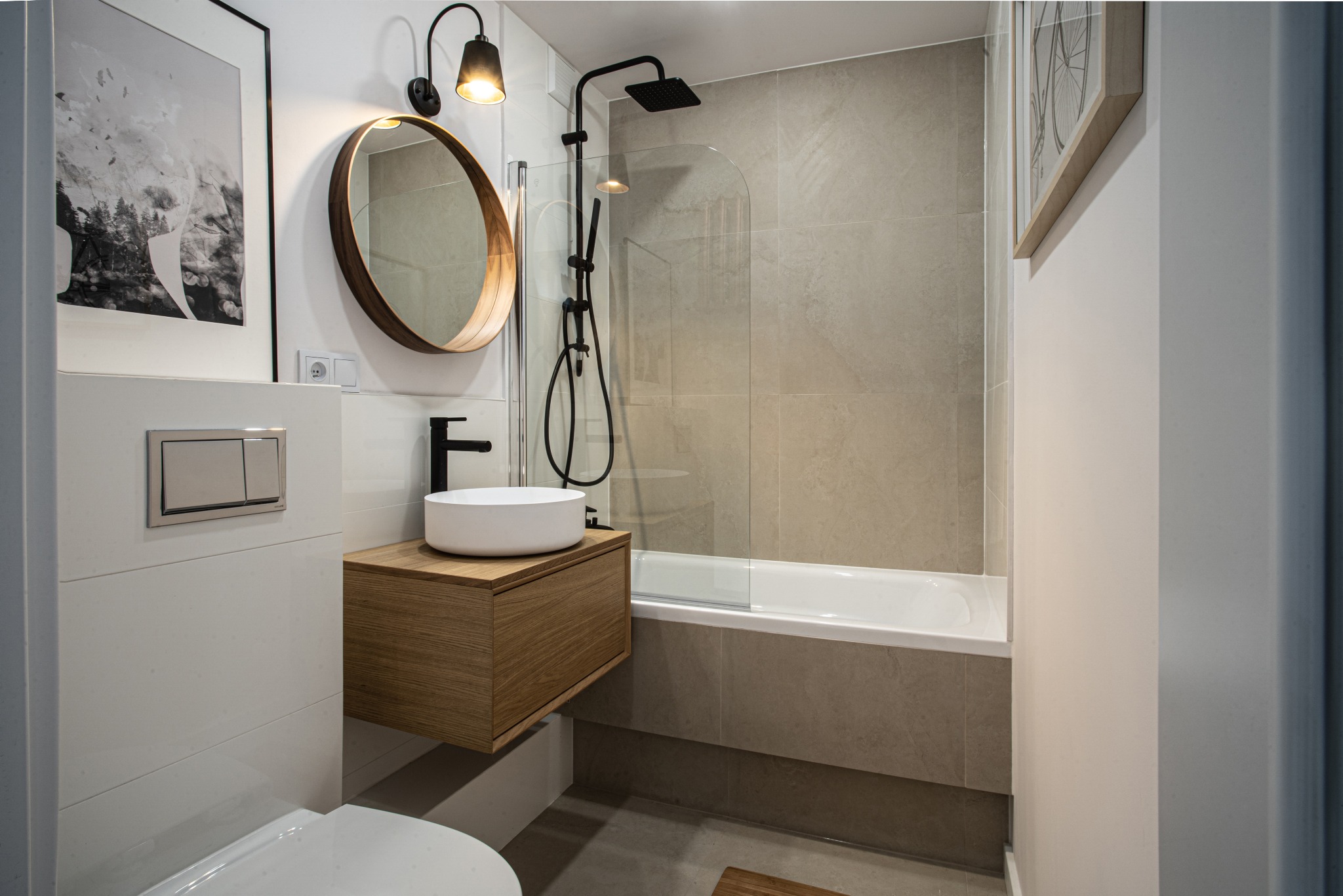 Nowoczesna łazienka z wiszącą szafką z umywalką, czarną armaturą, wanną z parawanem, okrągłym lustrem z drewnianą ramą i minimalistycznym oświetleniem.