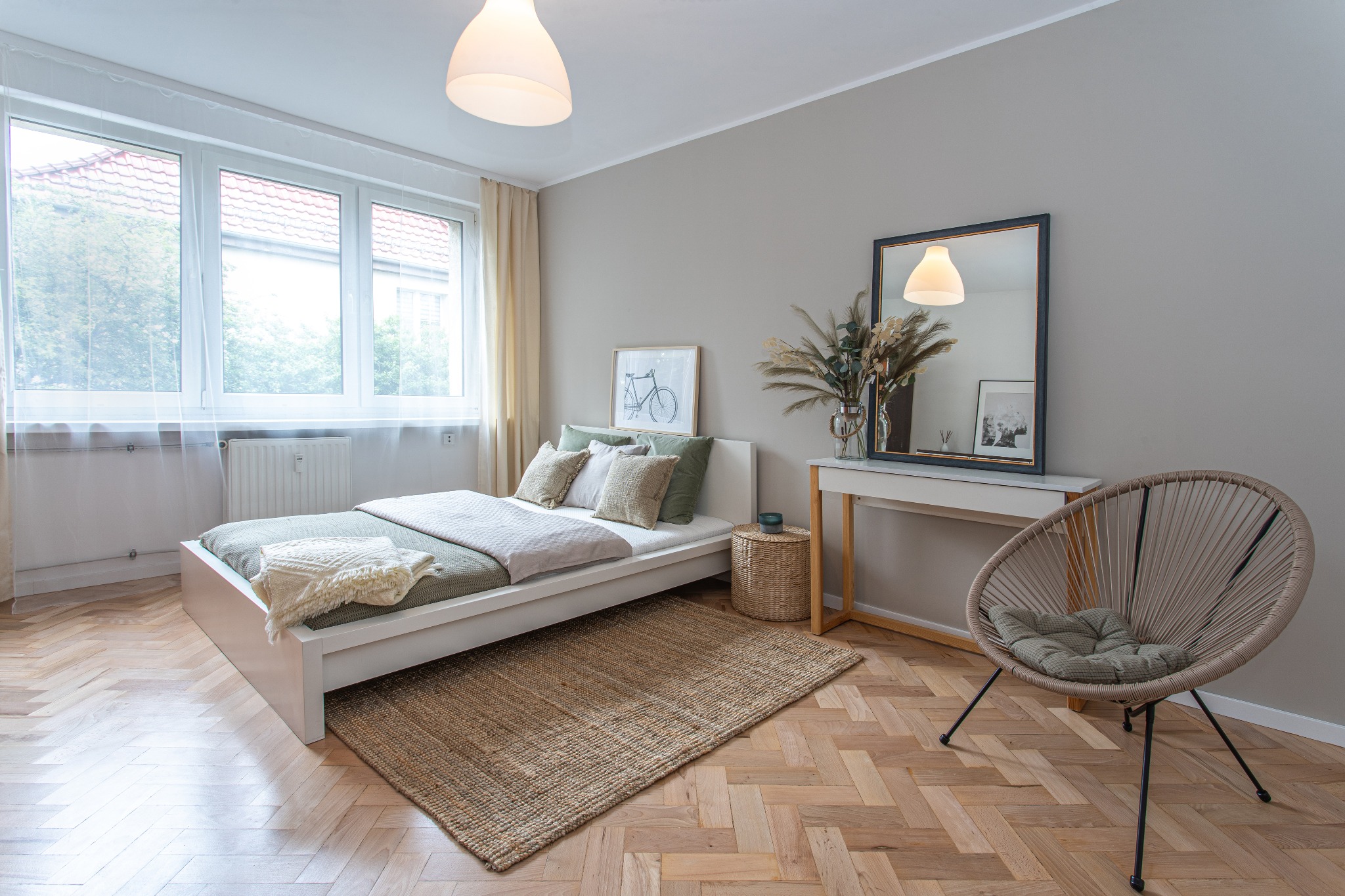 Jasna sypialnia z białym łóżkiem, drewnianą podłogą w jodełkę, jutowym dywanem, okrągłym krzesłem z poduszką i lustrem na konsoli.