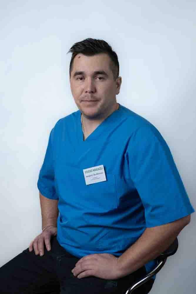 Portret mężczyzny w niebieskim uniformie z identyfikatorem 'Studio Masażu', siedzącego na krześle na tle białej ściany.