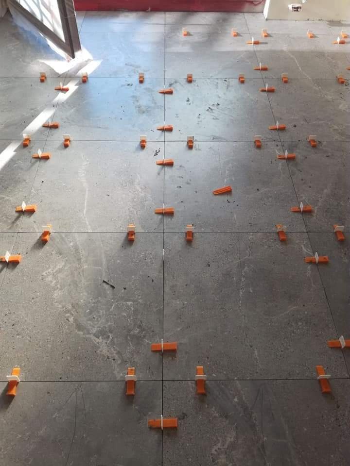 Szare płytki podłogowe w trakcie układania, z pomarańczowymi klinami poziomującymi w spoinach, widoczne linie fug.