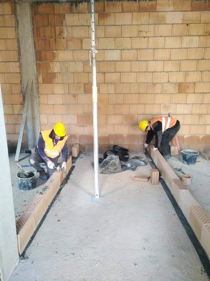 Dwóch robotników w żółtych kaskach i kamizelkach układa pustaki ceramiczne na zaprawie, tworząc fundament pod ścianę w surowym wnętrzu budynku. Widoczna stalowa podpórka stropowa i ceglana ściana...