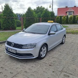 Srebrny Volkswagen Jetta z żółtym znakiem TAXI na dachu, zaparkowany na betonowym placu z widocznym numerem rejestracyjnym GWE 95278, w tle żywopłot z tuj i budynek z czerwoną dachówką.