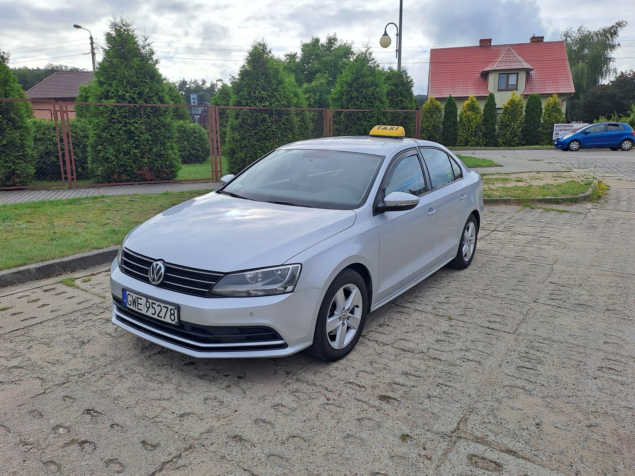 Srebrny Volkswagen Jetta z żółtym znakiem TAXI na dachu, zaparkowany na betonowym placu z widocznym numerem rejestracyjnym GWE 95278, w tle żywopłot z tuj i budynek z czerwoną dachówką.