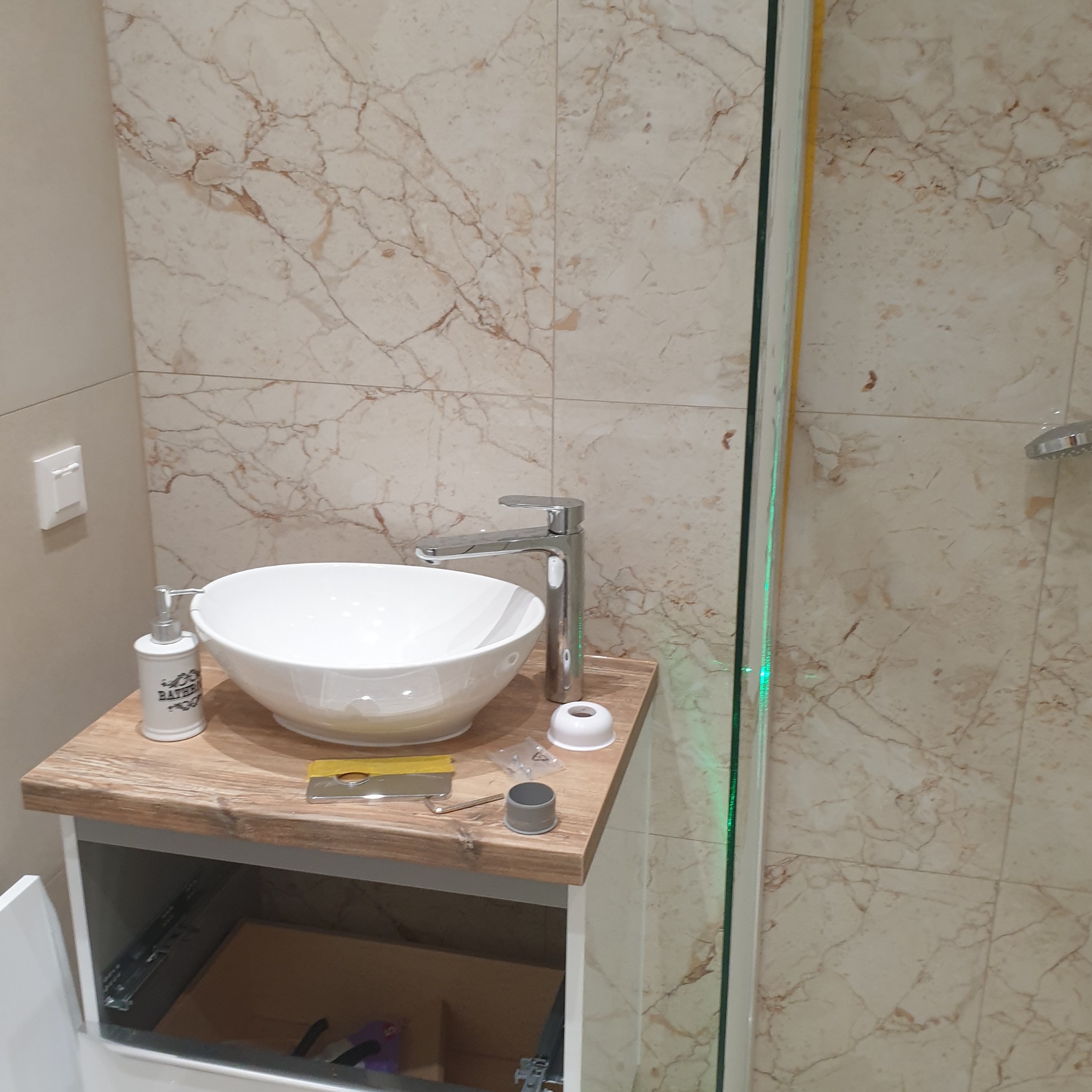 Montaż umywalki nablatowej w łazience z marmurowymi płytkami, widoczne narzędzia i elementy instalacyjne, szafka pod umywalką z otwartymi drzwiczkami.