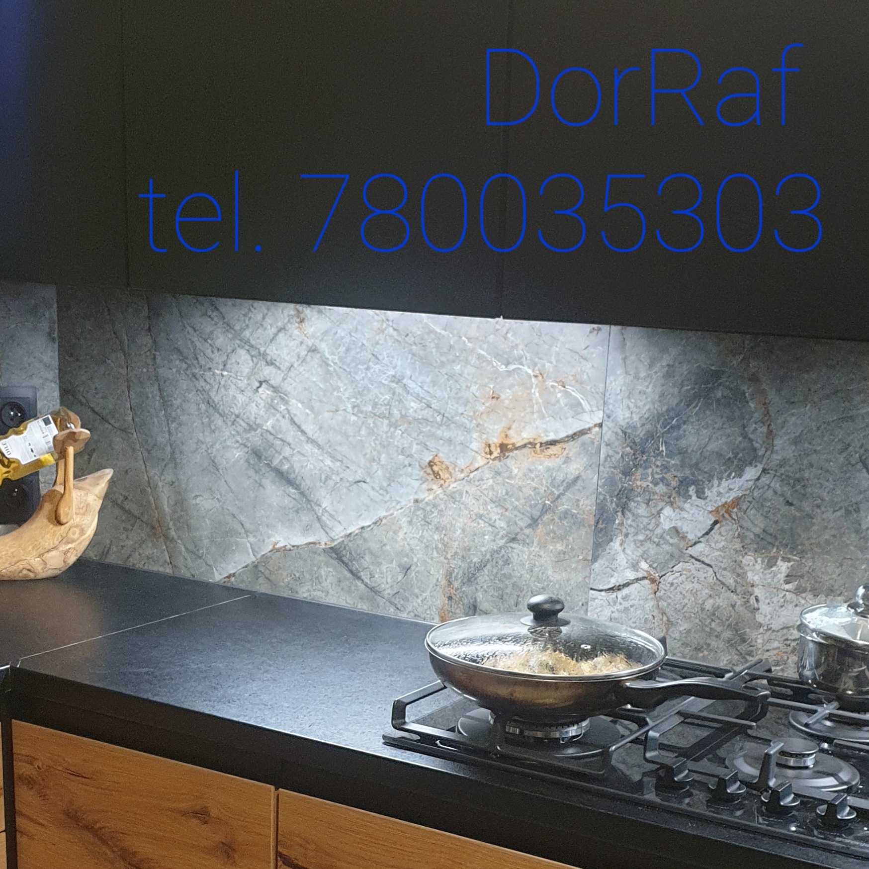Częściowy widok kuchni z marmurowym panelem ściennym, czarnym blatem, drewnianymi szafkami i czarną płytą grzewczą z dwoma garnkami. Na ścianie widoczny niebieski napis 'DorRaf tel. 780035303'.