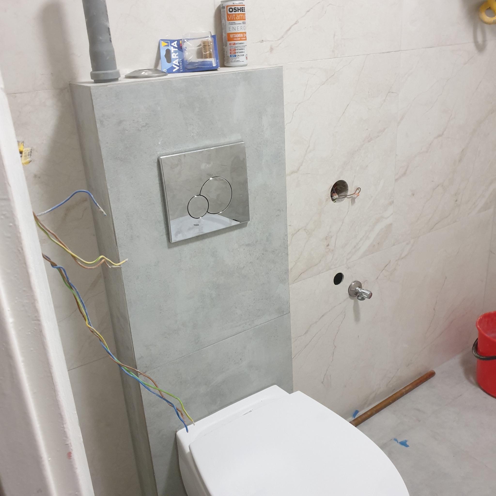 Podtynkowa instalacja toalety w trakcie prac wykończeniowych, widoczne wystające przewody elektryczne, płytki imitujące marmur oraz puszka z witaminami na zabudowie.