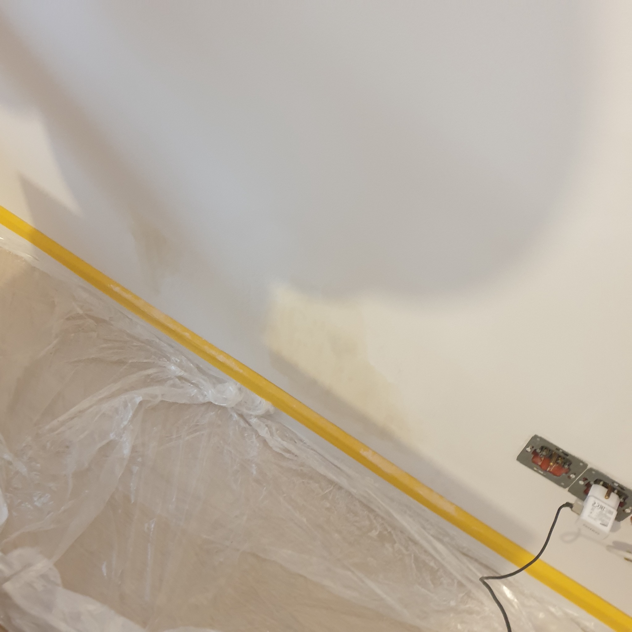 Przygotowana do malowania ściana z żółtą taśmą malarską przy podłodze zabezpieczonej folią, widoczne gniazdka elektryczne z podłączonym zasilaczem.