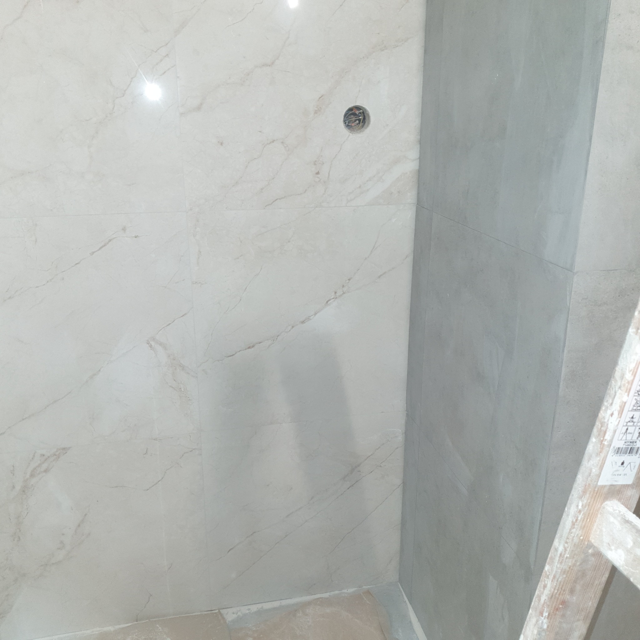 Ściana w trakcie wykańczania, częściowo pokryta jasnymi płytkami imitującymi marmur, z widocznym gniazdkiem instalacyjnym i szarą, gładką powierzchnią obok, fragment drabiny w prawym dolnym rogu.