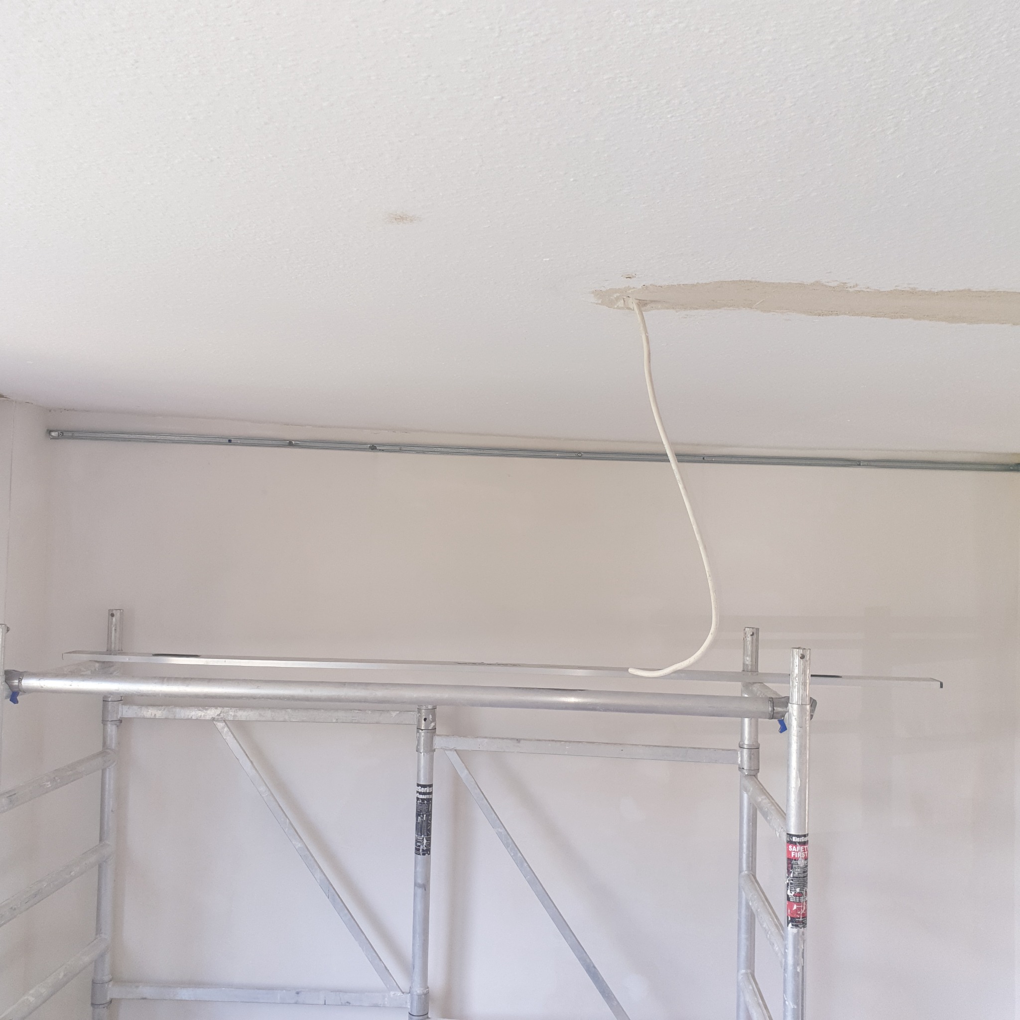 Wnętrze w trakcie prac wykończeniowych; widoczne rusztowanie, kabel wystający z sufitu oraz ślady szpachlowania.