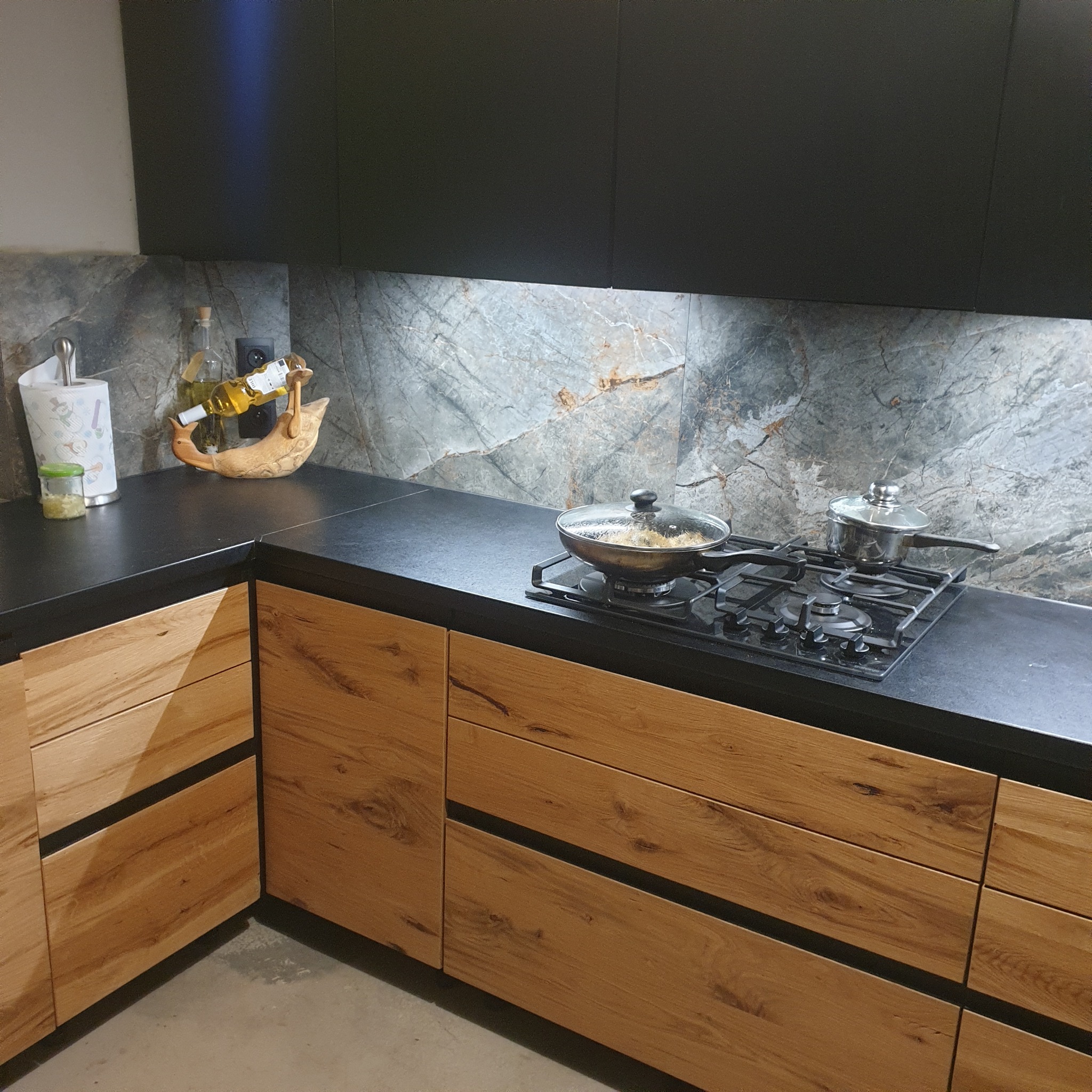 Nowoczesna kuchnia z drewnianymi frontami szafek i czarnym blatem, na którym stoi płyta gazowa z garnkiem i patelnią, a w tle marmurowa ściana.