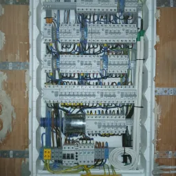 Rozdzielnica elektryczna w białej obudowie, zamontowana w surowej ścianie z cegły. Widoczne liczne przewody, zabezpieczenia i elementy instalacji.