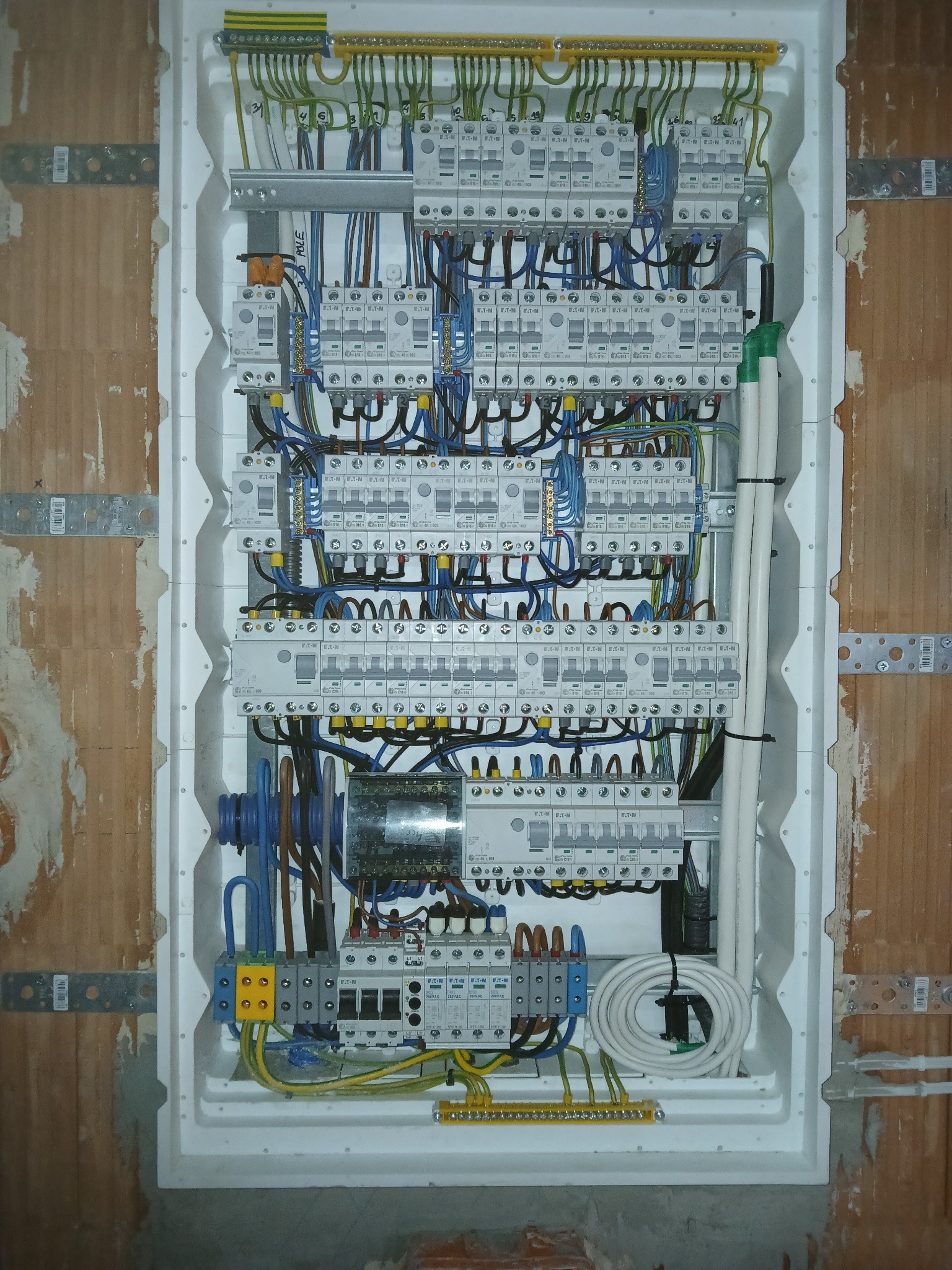 Rozdzielnica elektryczna w białej obudowie, zamontowana w surowej ścianie z cegły. Widoczne liczne przewody, zabezpieczenia i elementy instalacji.