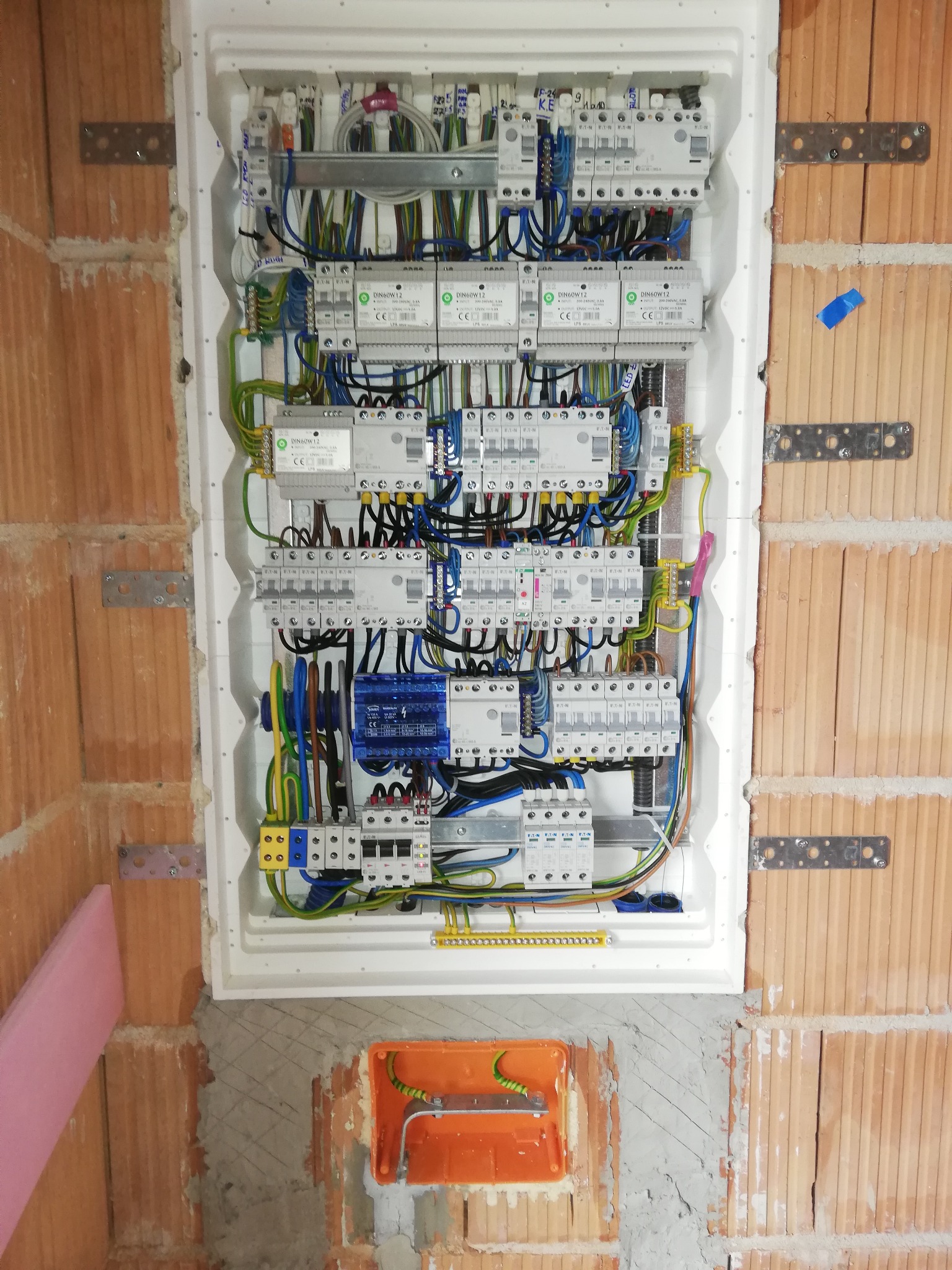 Rozdzielnica elektryczna w obudowie, zamontowana na ścianie z cegły. Widoczne zabezpieczenia, kable i przewody. Instalacja w trakcie montażu.