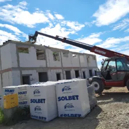 Czerwona ładowarka teleskopowa Manitou podnosi materiały budowlane na budowę dwukondygnacyjnego budynku z białych bloczków Solbet, widoczne palety z bloczkami, błękitne niebo z chmurami w tle.