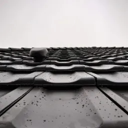 Czarny dach z dachówki ceramicznej, pokryty kroplami deszczu, z widocznym odpowietrznikiem dachowym na tle pochmurnego nieba, sfotografowany pod kątem od dołu.