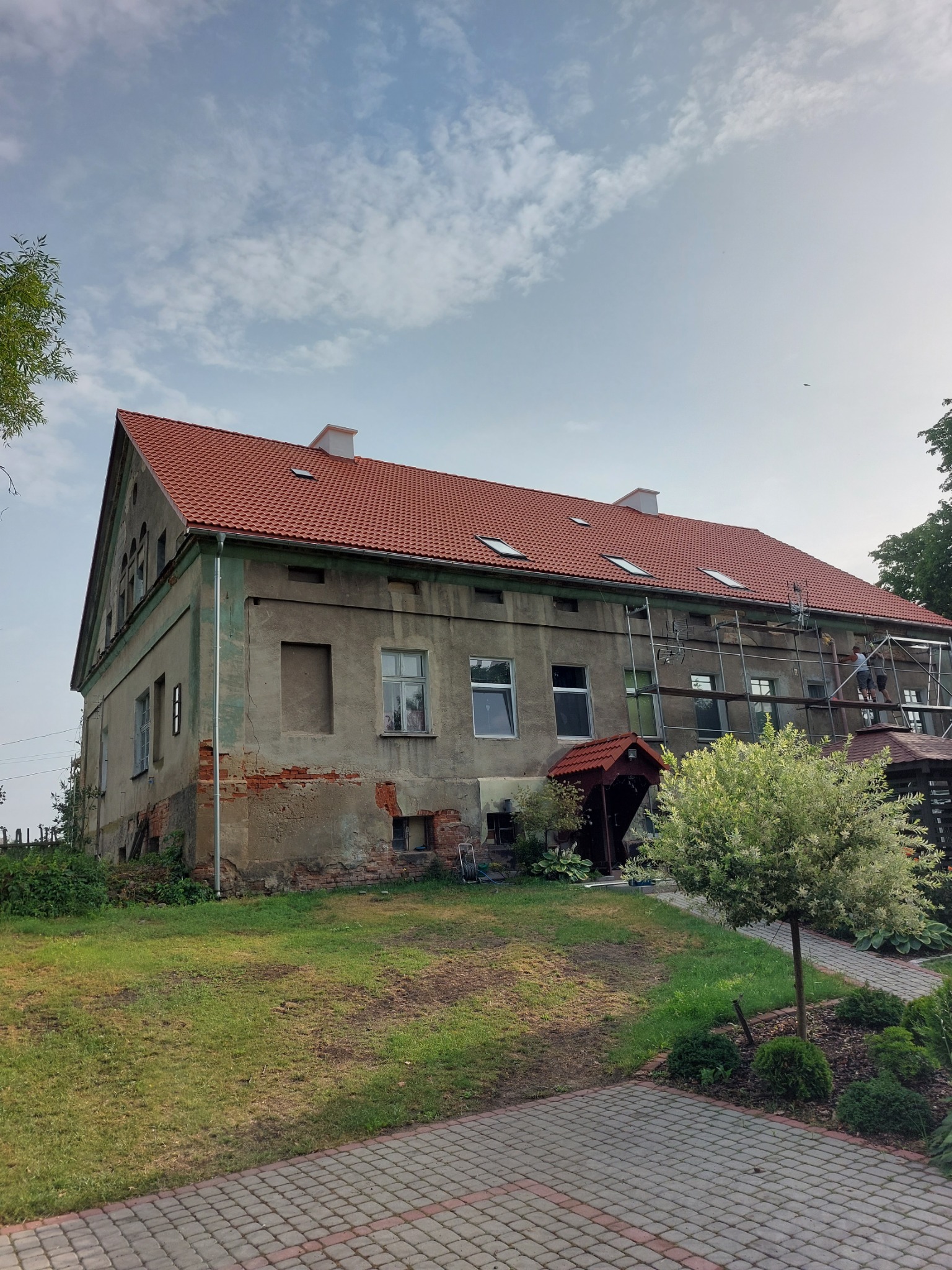 Dwupiętrowy budynek z czerwonym dachem, częściowo odnowiony, z rusztowaniem i pracownikami na rusztowaniu, otoczony zielonym trawnikiem i kostką brukową.