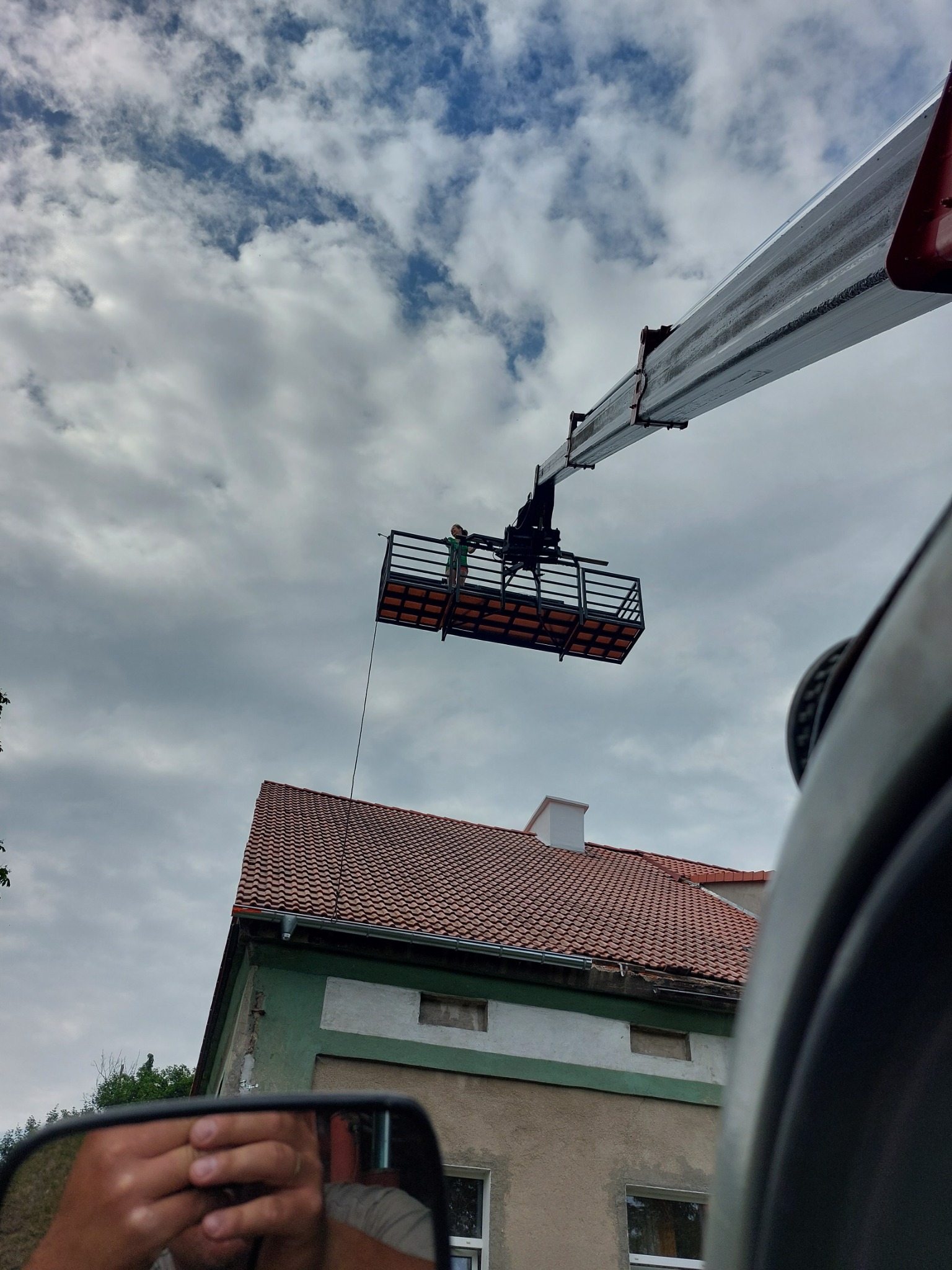 Pracownik w koszu ładowarki teleskopowej na tle pochmurnego nieba, widoczny dach budynku z czerwonej dachówki i fragment samochodu z lusterkiem wstecznym.
