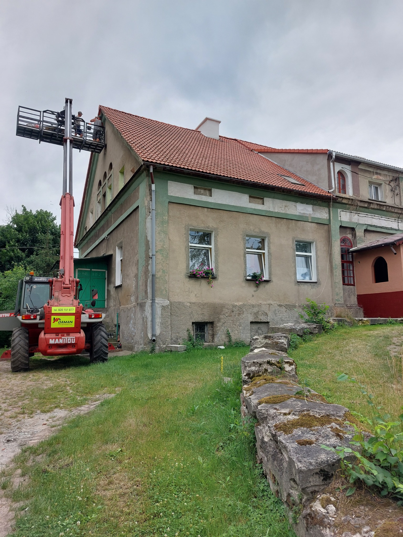 Dwóch pracowników na podnośniku teleskopowym MANITOU, pracujących przy dachu starego, dwupiętrowego budynku z czerwoną dachówką. Na ścianie budynku widoczne okna z kwiatami.
