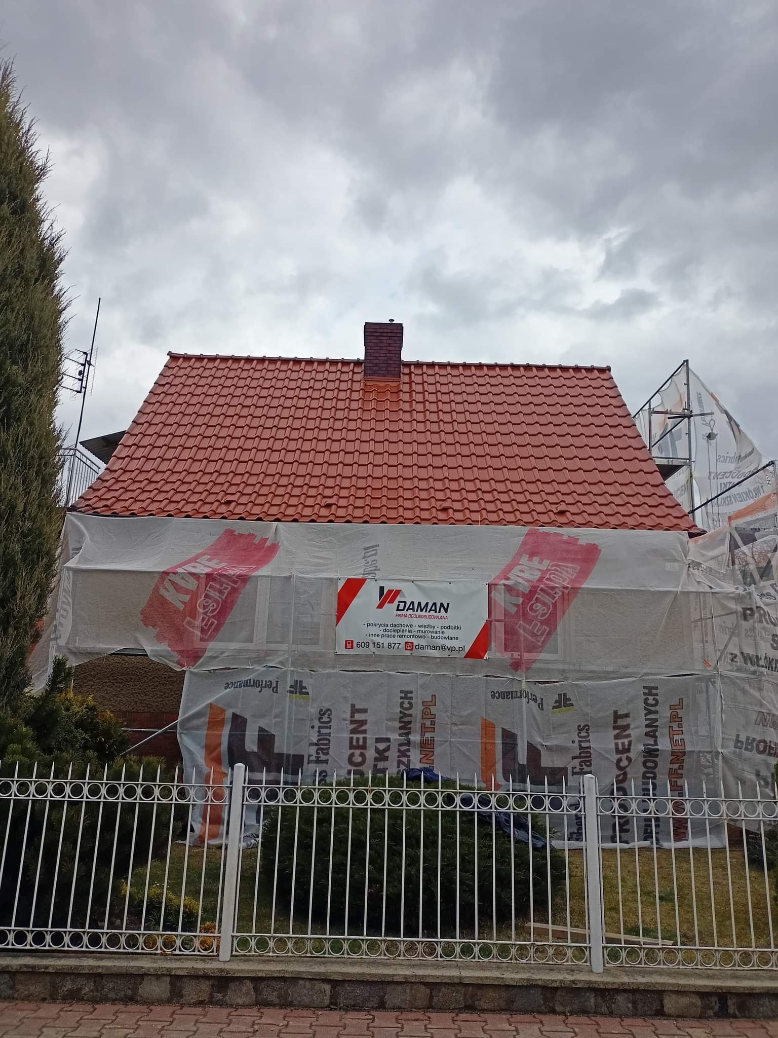 Dom z nowym, czerwonym dachem z dachówki, częściowo osłonięty folią z reklamą firmy budowlanej DAMAN, widoczny zza białego, metalowego ogrodzenia w pochmurny dzień.