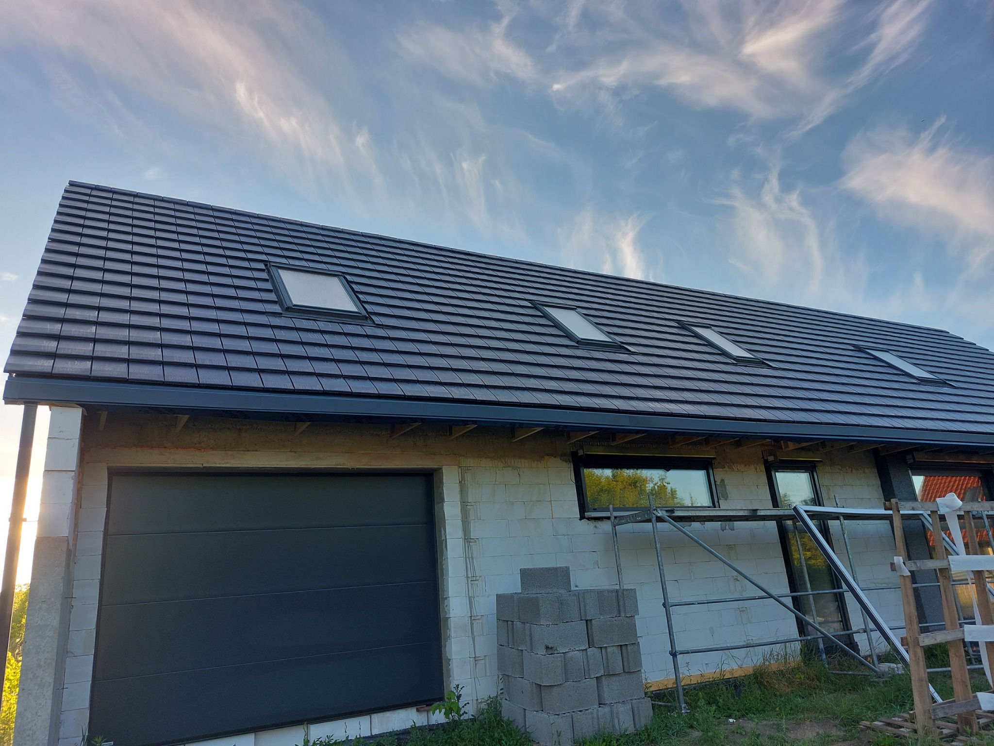 Nowy, grafitowy dach z czterema oknami połaciowymi na surowym budynku z białego gazobetonu, widoczne rusztowania i składowane bloczki.