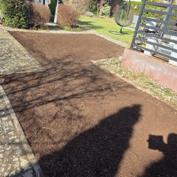 Marek przybył - Przygotowany teren pod trawnik z widoczną strukturą gleby, obok ścieżka z kostki brukowej i elementy architektury ogrodowej w tle. Cień osoby robiącej zdjęcie.