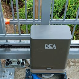 SPIDPOOL Paweł Łukaszyk - Zamontowany napęd DEA przy bramie przesuwnej w Zamościu. Widoczna listwa zębata i elementy mocujące. Ujęcie z bliska, zieleń w tle.