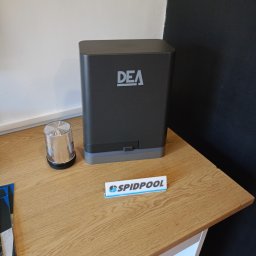 SPIDPOOL Paweł Łukaszyk - Szary napęd DEA z lampą ostrzegawczą i wizytówką na drewnianym blacie, na tle białej i czarnej ściany. Widoczny fragment podłogi z terakoty.