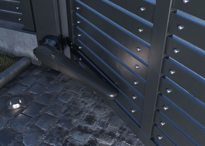 Szara, metalowa brama z automatycznym napędem, widoczny mechanizm siłownika na tle kostki brukowej i wbudowanego oświetlenia.