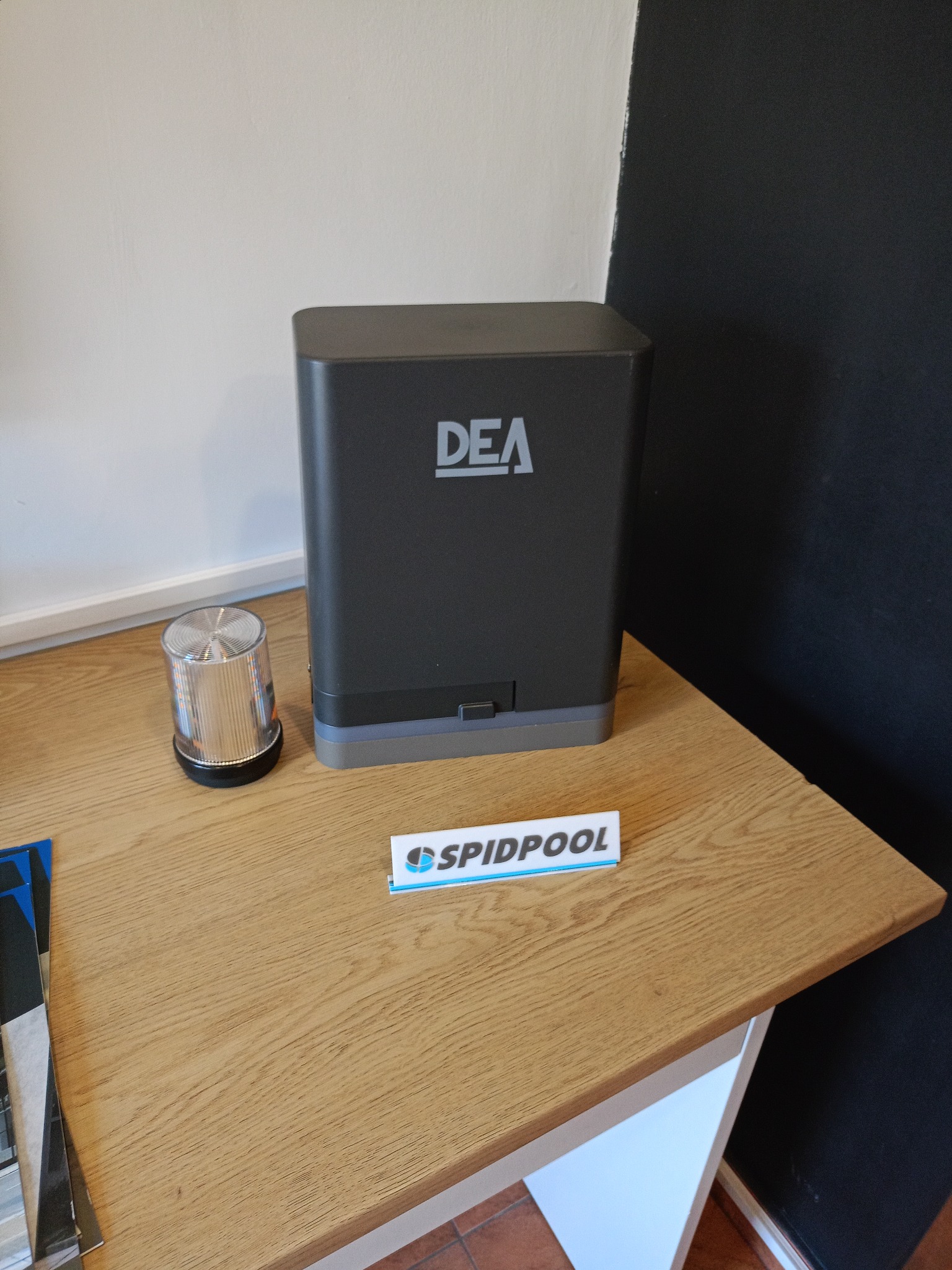 Szary napęd DEA z lampą ostrzegawczą i wizytówką na drewnianym blacie, na tle białej i czarnej ściany. Widoczny fragment podłogi z terakoty.