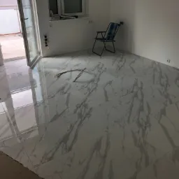 Pokój z nowo położoną, błyszczącą podłogą z białych płytek imitujących marmur; widoczne kable elektryczne na podłodze, otwarte drzwi balkonowe i krzesło turystyczne w rogu.