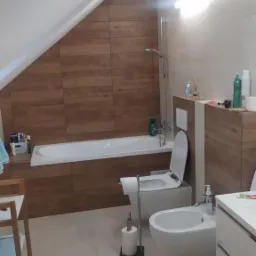 Wykończona łazienka na poddaszu z drewnopodobnymi płytkami na ścianach, białą wanną, toaletą i bidetem. Widoczny stojak na papier toaletowy i biała szafka umywalkowa.