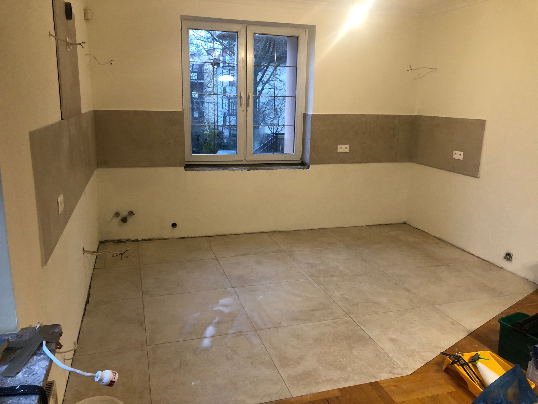 Wnętrze kuchni w trakcie remontu, widoczne świeżo położone płytki podłogowe w odcieniach beżu, częściowo ułożone płytki ścienne, odsłonięte przewody elektryczne i narzędzia pracy w rogu pomieszczenia.