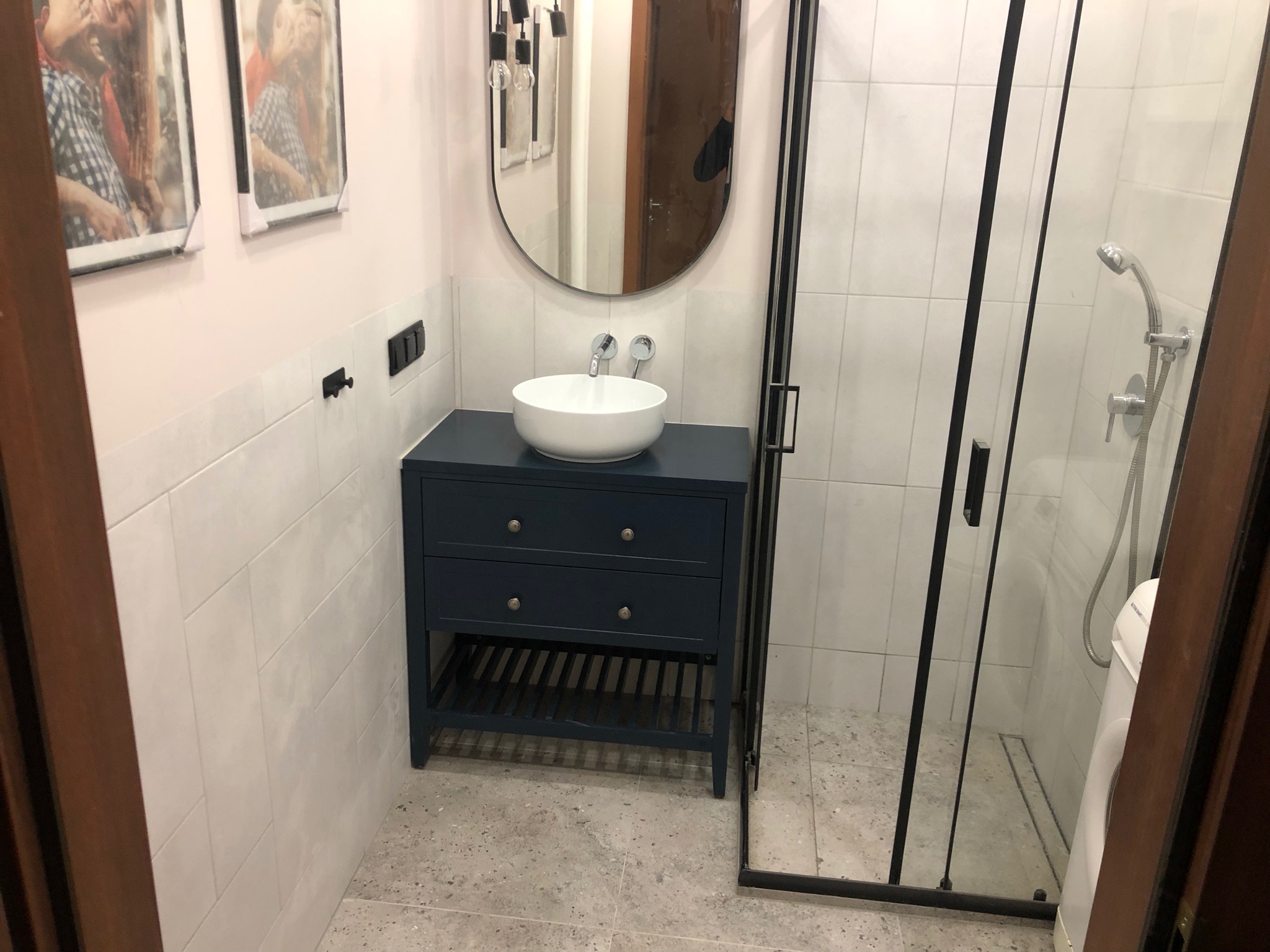 Nowoczesna łazienka z kabiną prysznicową w czarnej ramie, białą umywalką nablatową na granatowej szafce z dwoma szufladami i owalnym lustrem bez ramy, płytki imitujące kamień na podłodze i ścianach.