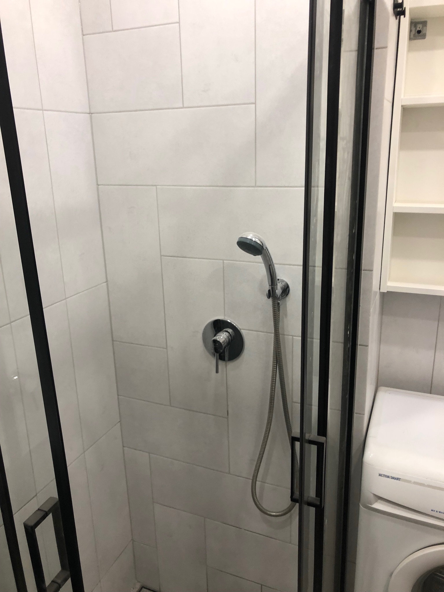 Kabina prysznicowa z czarnymi profilami, białe, prostokątne płytki na ścianach, bateria z deszczownicą, pralka w tle, szafka wisząca otwarta.