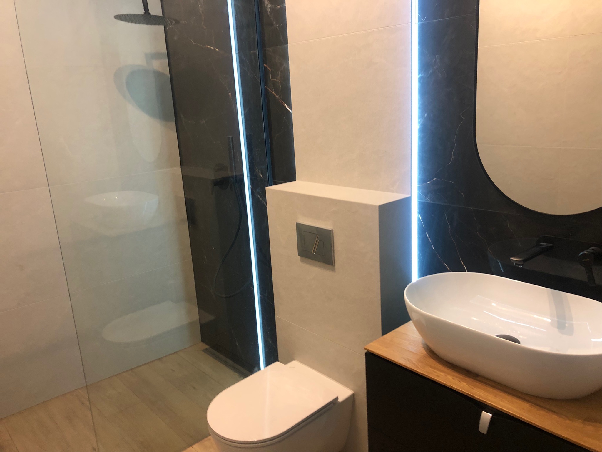 Nowoczesna łazienka z kabiną prysznicową typu walk-in, umywalką nablatową na drewnianym blacie i podświetleniem LED przy czarnych płytkach imitujących marmur.