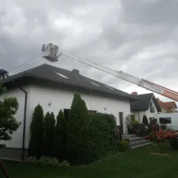 Prace dekarskie przy użyciu podnośnika koszowego na tle pochmurnego nieba, widoczny fragment dachu z dachówką ceramiczną i pracownicy w koszu oraz na dachu.