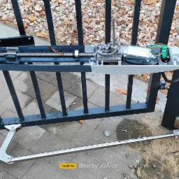 Widok z bliska na zamontowany mechanizm automatyki bramy skrzydłowej, z widocznymi elementami silnika, przekładni ślimakowej i ramienia teleskopowego, na tle kostki brukowej i opadłych liści.