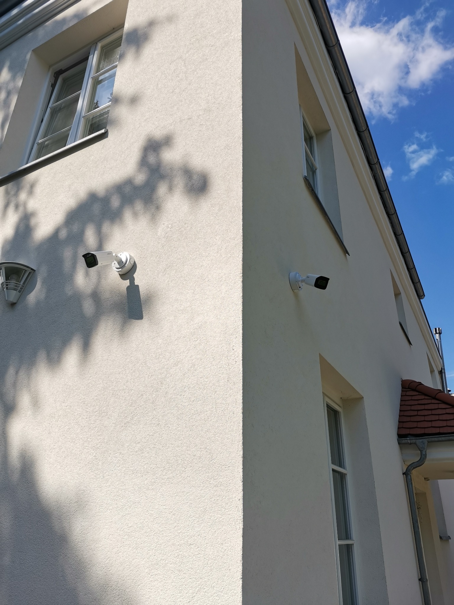 Dwie białe kamery monitoringu zamontowane na elewacji budynku z jasną, chropowatą fakturą, widoczne okna i fragment dachu z czerwoną dachówką, cień drzewa na ścianie, błękitne niebo w tle.