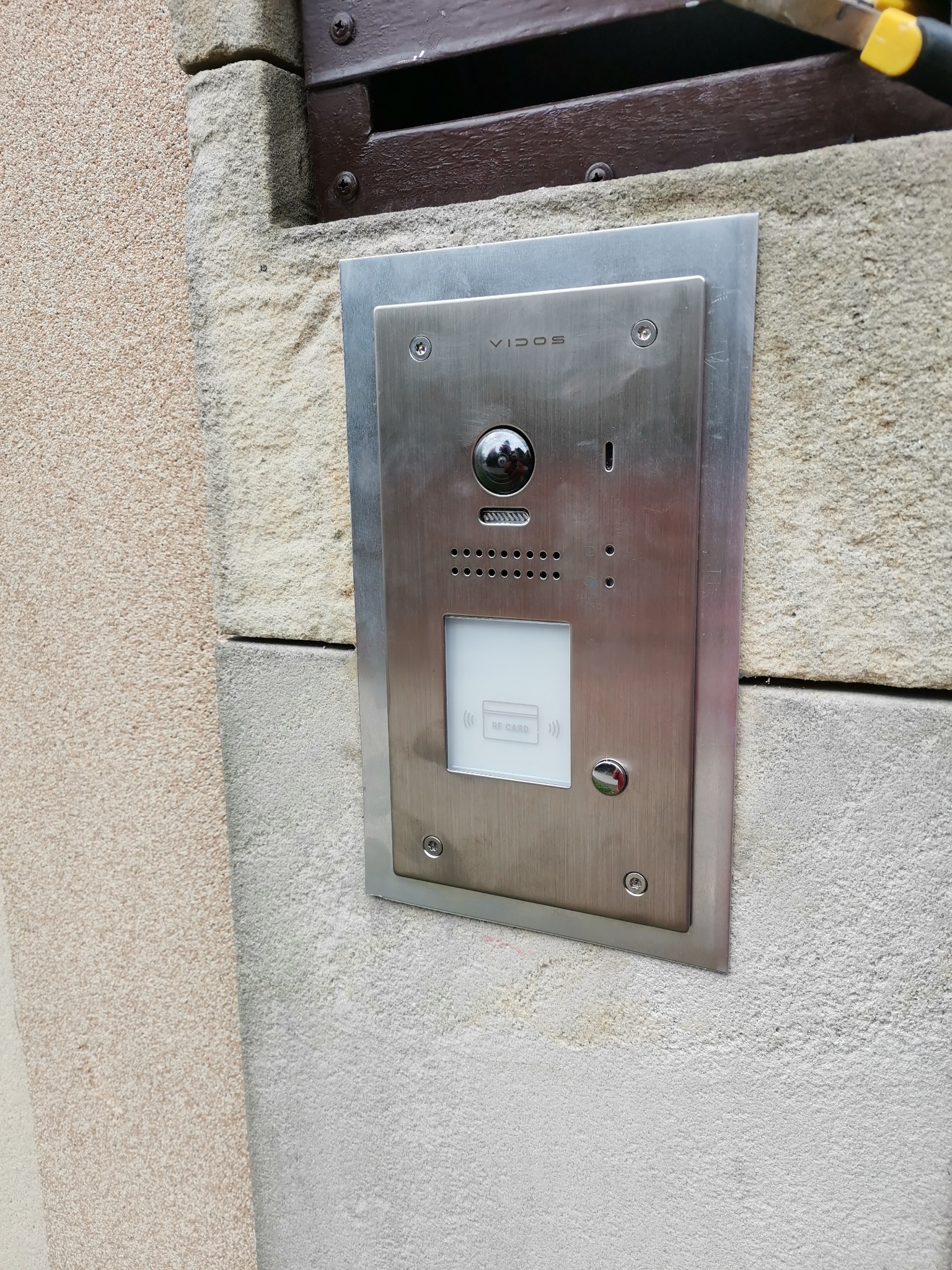 Zewnętrzny panel domofonowy Vidos z kamerą i czytnikiem kart zbliżeniowych, zamontowany na nierównej, jasnej elewacji budynku.