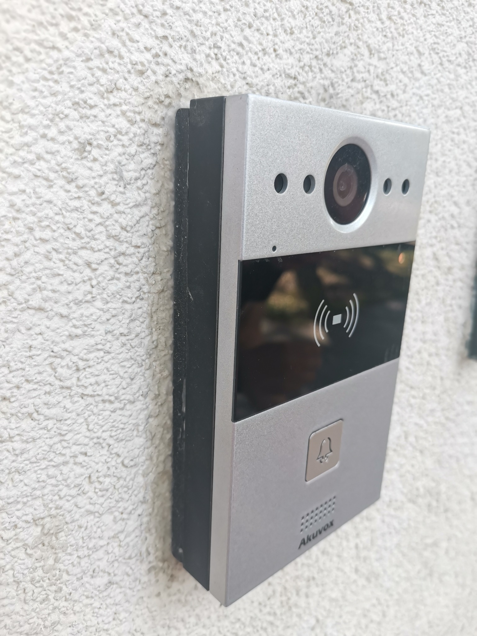 Nowoczesny, srebrny wideodomofon Akuvox z czytnikiem kart zbliżeniowych, zamontowany na białej, chropowatej ścianie zewnętrznej budynku.