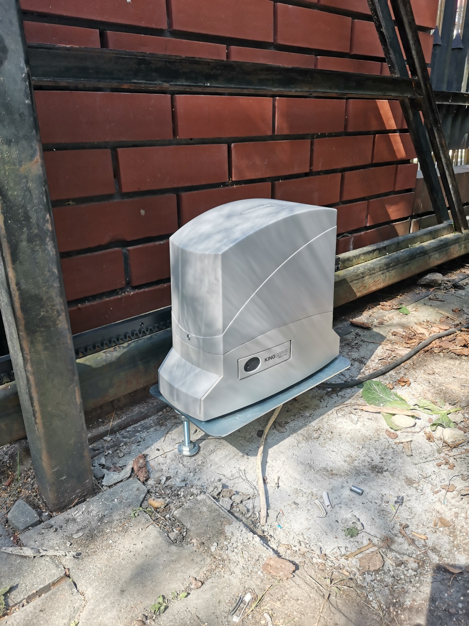 Szary napęd do bram przesuwnych King Gates zamontowany na metalowej podstawie przy ceglanym murze i szynie bramy, widoczne ślady montażu na betonowej nawierzchni.