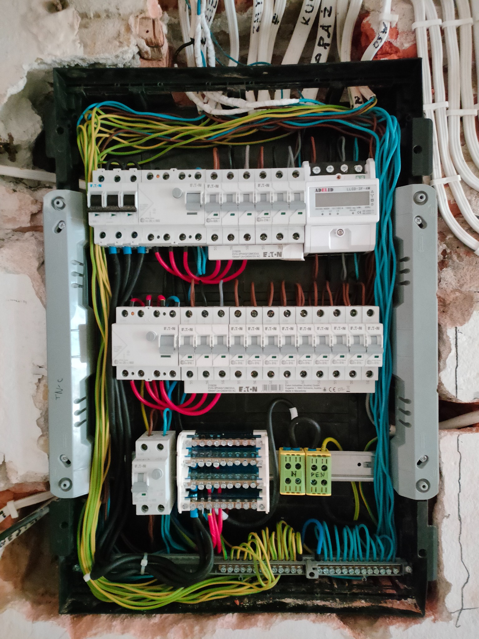 Otwarta skrzynka elektryczna z widocznymi bezpiecznikami, przewodami w różnych kolorach, listwami zaciskowymi i licznikiem energii, zamontowana w ścianie z widocznymi śladami po kuciu.