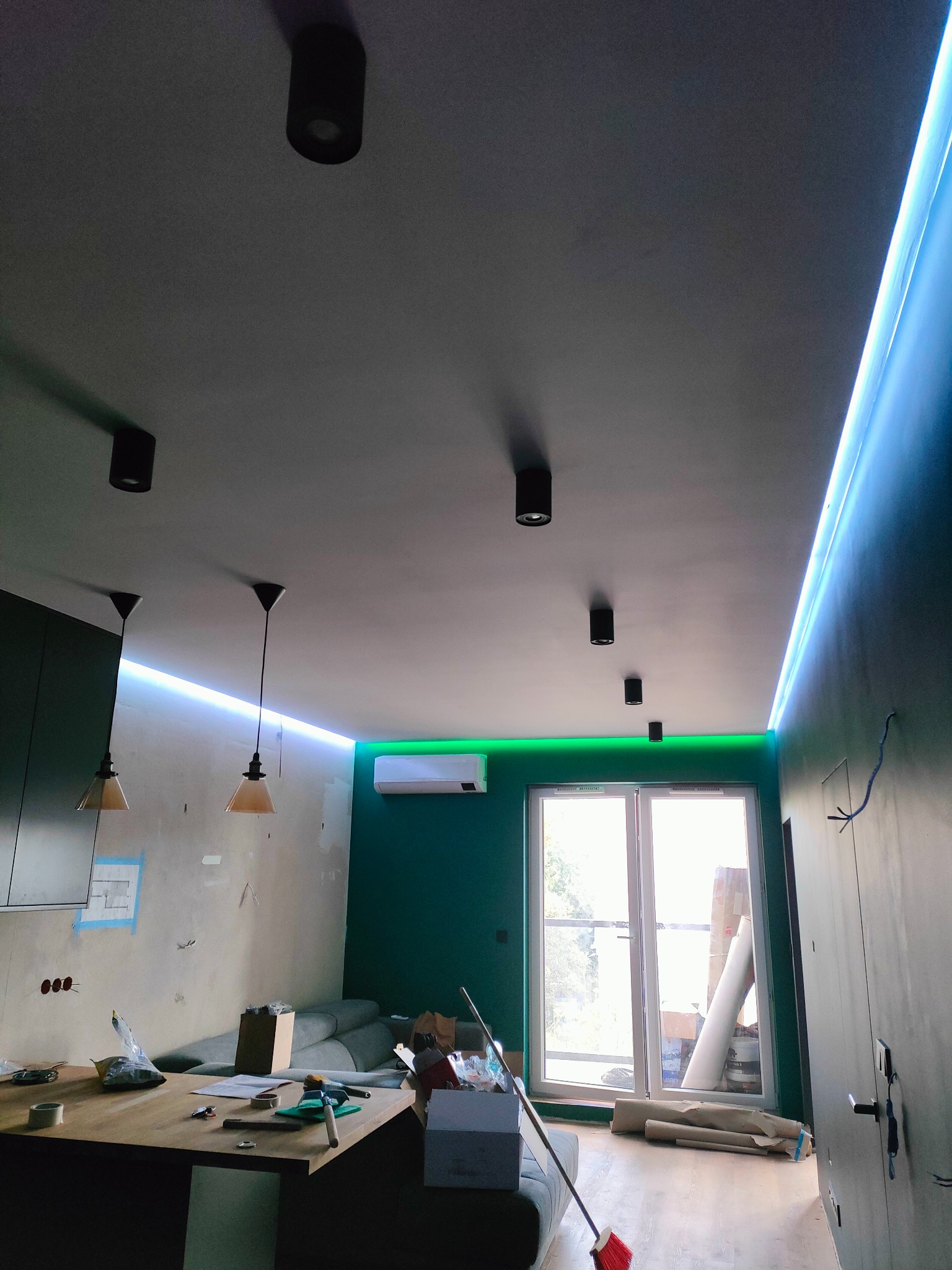 Wnętrze w trakcie remontu z widocznymi punktowymi lampami sufitowymi, podwieszonymi lampami z kloszami, paskiem LED na suficie oraz instalacją elektryczną w ścianie.