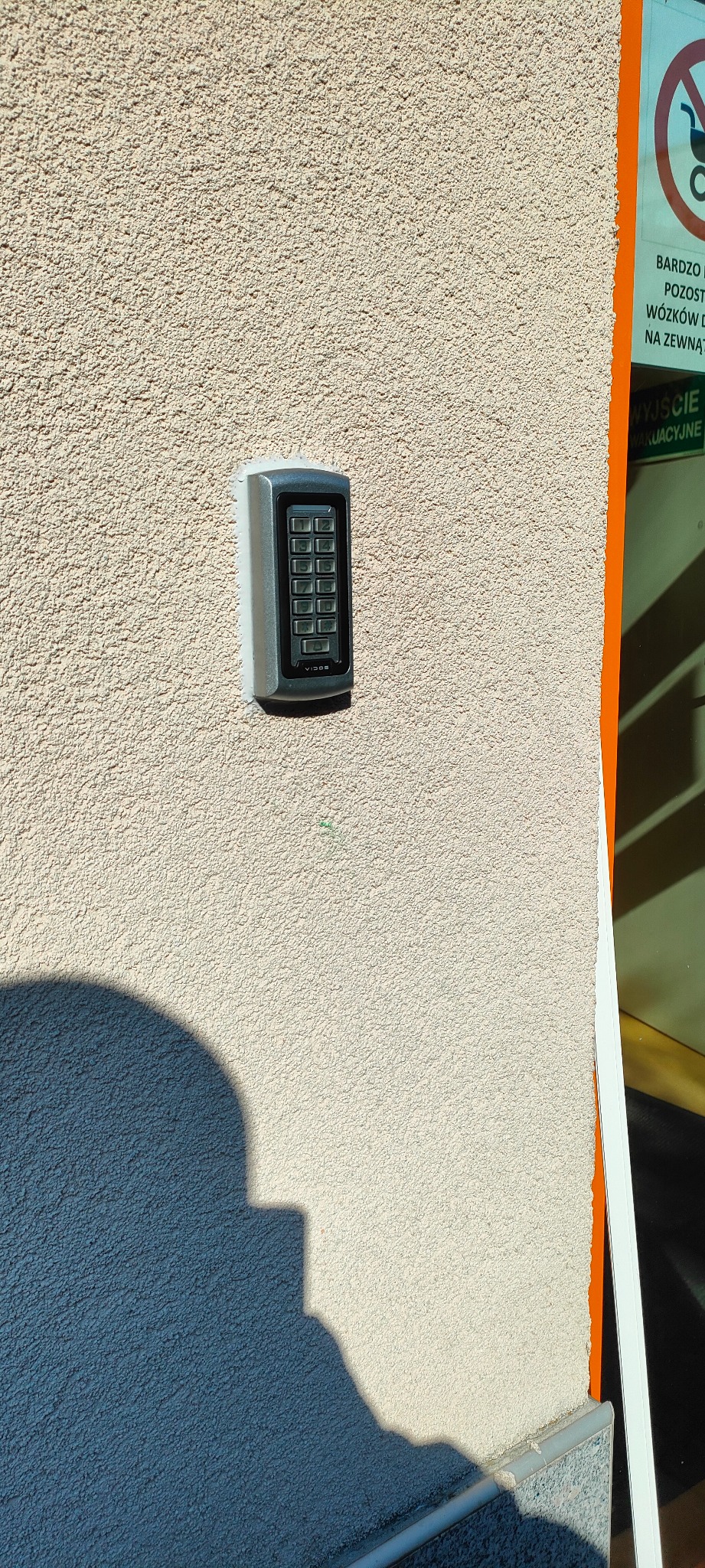 Szary panel numeryczny domofonu Vidos zamontowany na teksturowanej, beżowej ścianie budynku, obok pomarańczowej ramy drzwi wejściowych i cienia