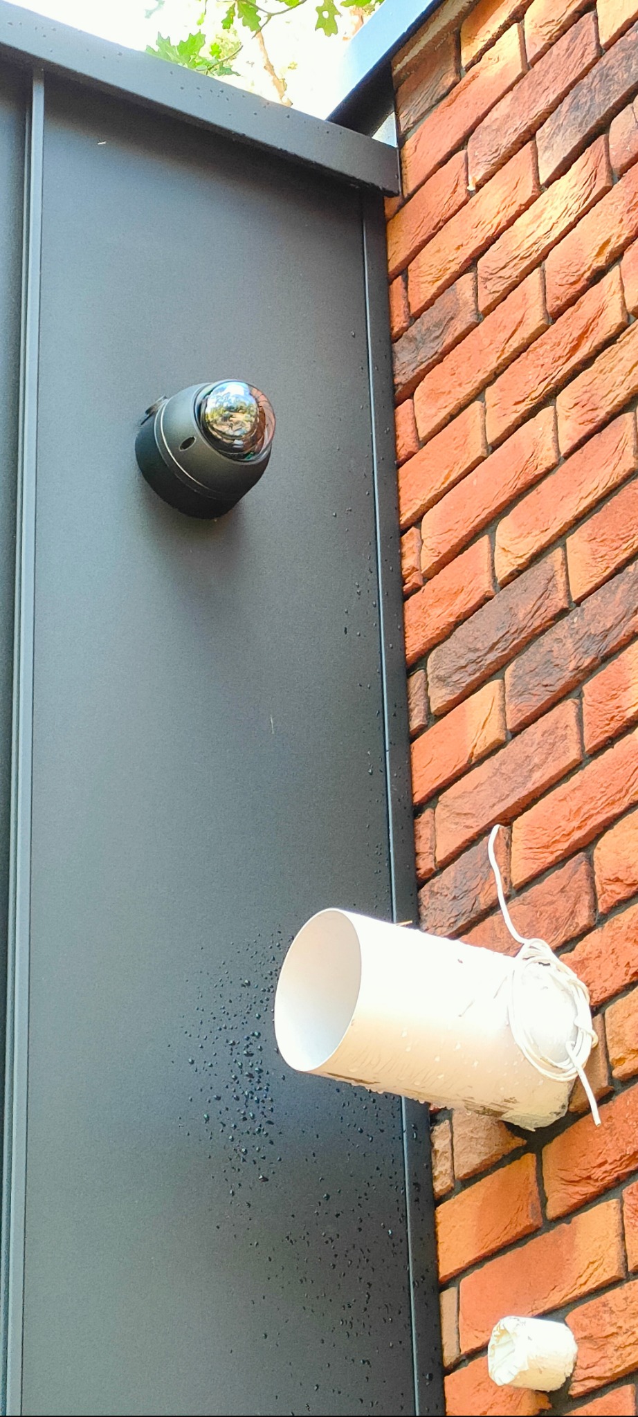 Kamera kopułkowa zamontowana na elewacji obok rury osłonowej z kablem, na tle ceglanej ściany i czarnej blachy.